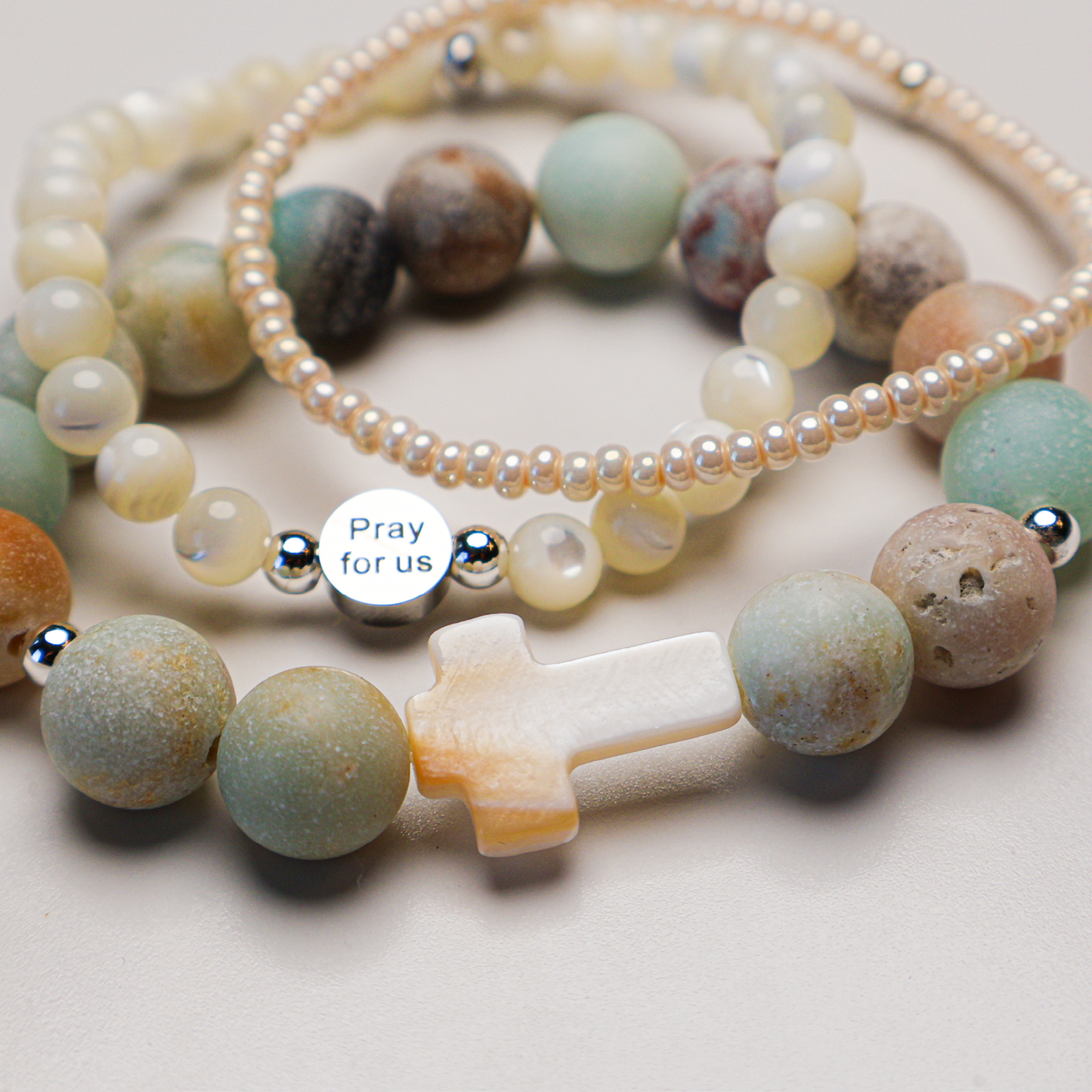 A set of three bracelets featuring a white stone cross against a beaded pattern, one with white beads, one with various colorful beads, and then a simple pearl-like complementary bracelet.